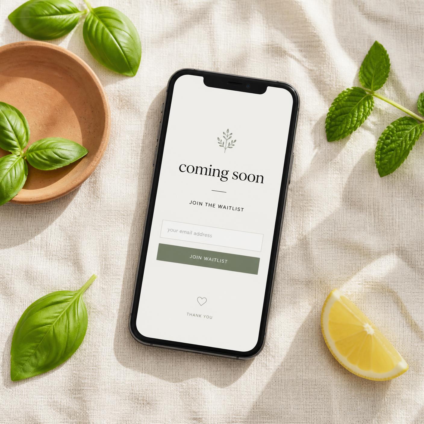 A phone on a linen cloth showing a coming soon waitlist screen, surrounded by fresh herbs